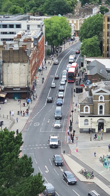 An aerial view of a city street during daytime showing a line of cars waiting in traffic, including a white van and a black car at the front, with pedestrians walking along the sidewalks. The street i