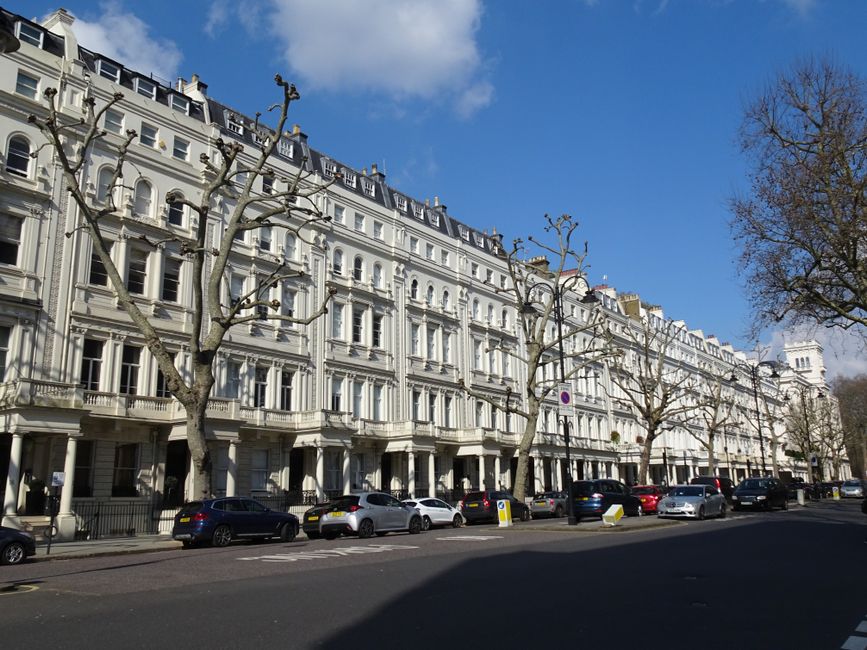 An aerial view of a city street during daytime showing a line of cars waiting in traffic, including a white van and a black car at the front, with pedestrians walking along the sidewalks. The street i